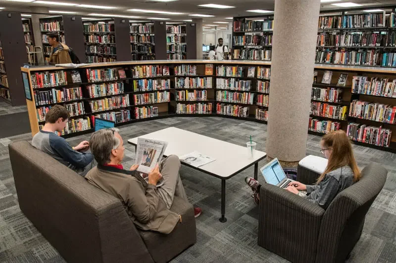 Personas leyendo y estudiando en una sala de biblioteca rodeada de estanterías con libros y revistas